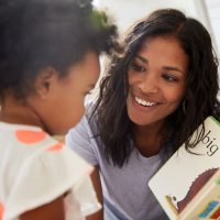 Woman with dark hair and skin reading book to a toddler