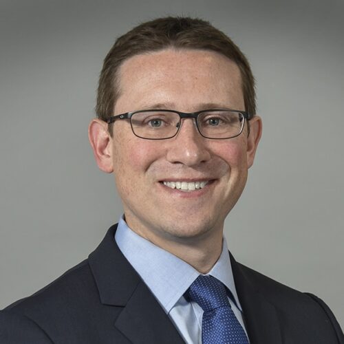 Smiling man in a suit, blue shirt, and glasses, posed against a plain gray background.