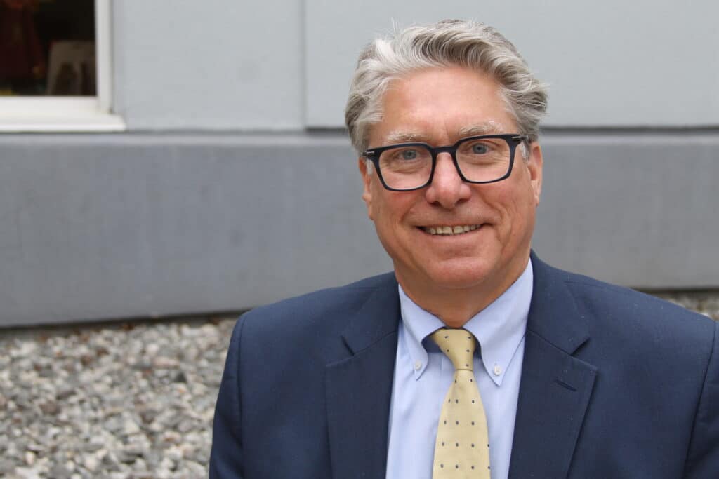 Smiling man in a suit and glasses stands outside against a gray wall and gravel ground.