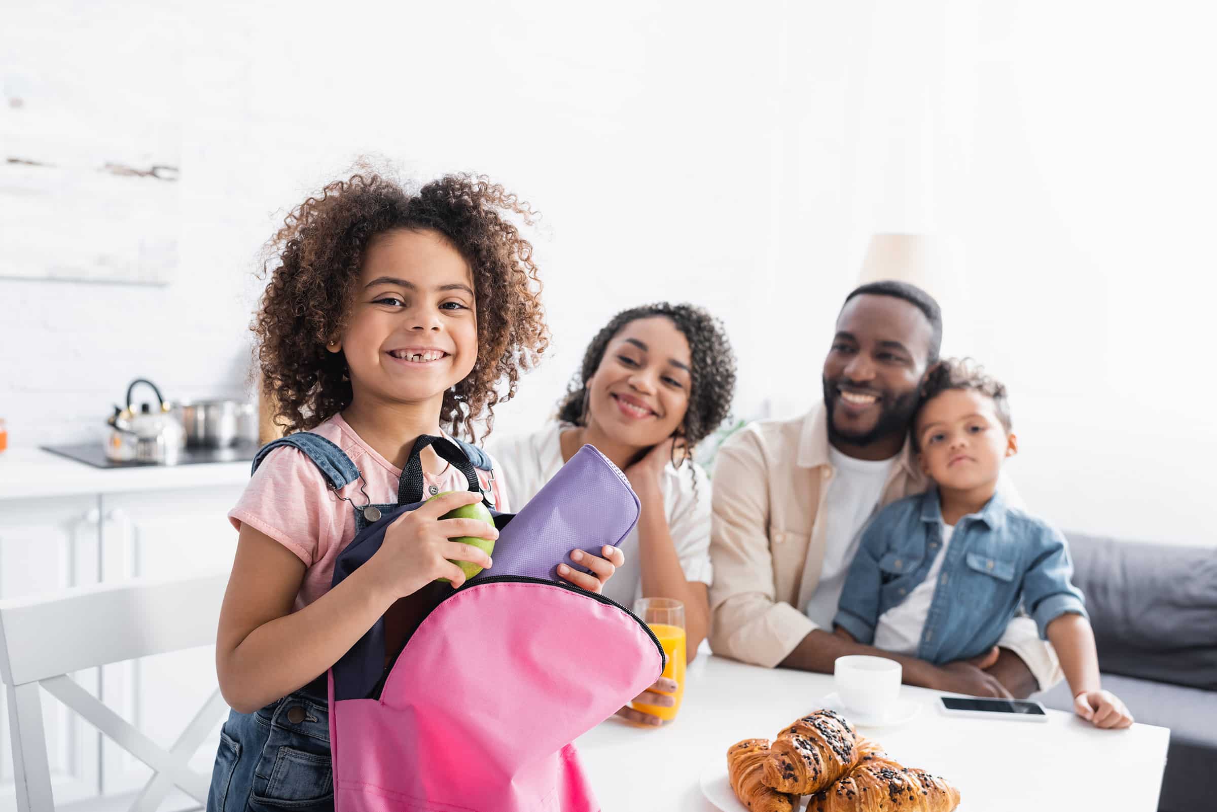 Smiling girl with a backpack stands in front of her cheerful family at a breakfast table.