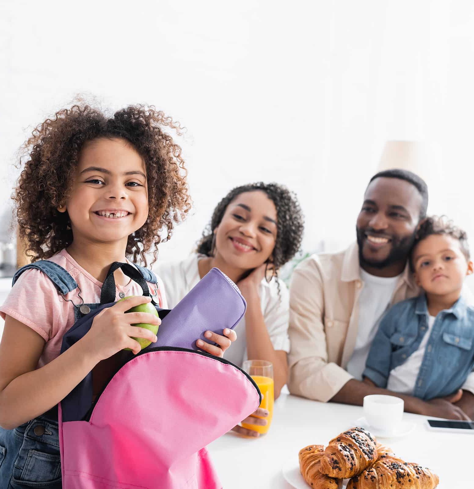 Young girl smiling at camera while holding backpack with parents and younger brother in background