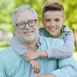 Older man with glasses and white hair with young boy, 12, with arms around neck and shoulders smiling at camera