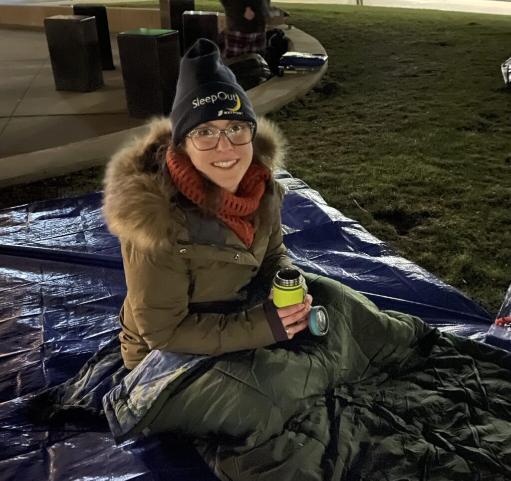 Young woman with hat and coat on in sleeping bag lying on top of tarp outside