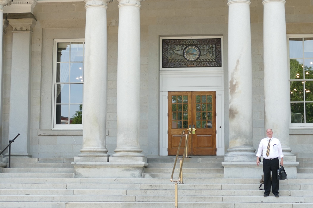 man with white shirt and black pants with briefcase standing on steps of government building