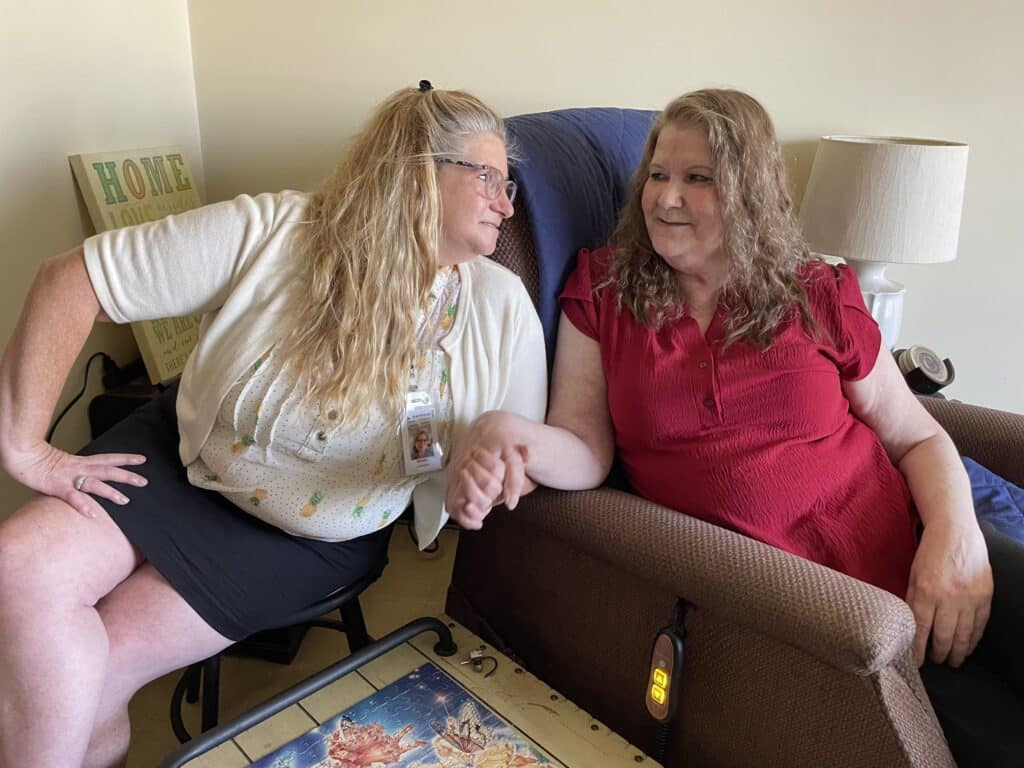 Two women sit close together, smiling and holding hands in a warmly lit living room.