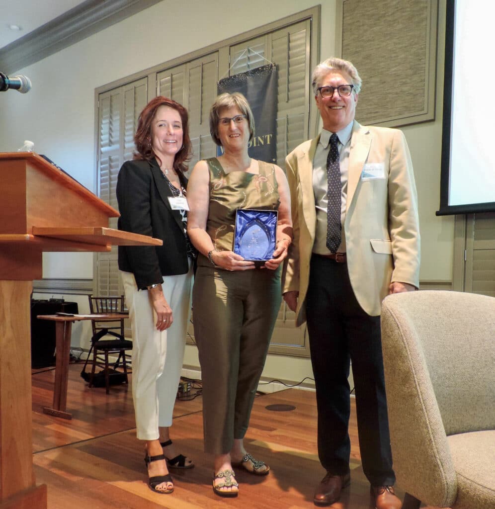 Three people stand indoors; the woman in the center holds a glass award and smiles at the camera.