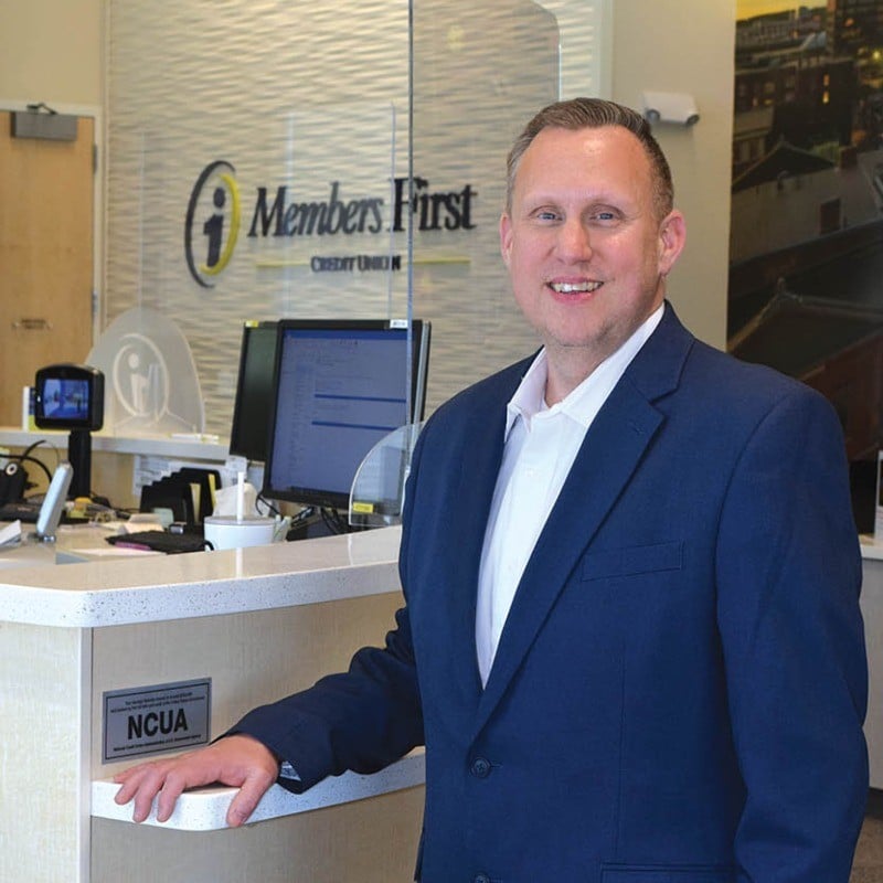 Man in a blue blazer stands smiling at a credit union counter with Members First sign in background.