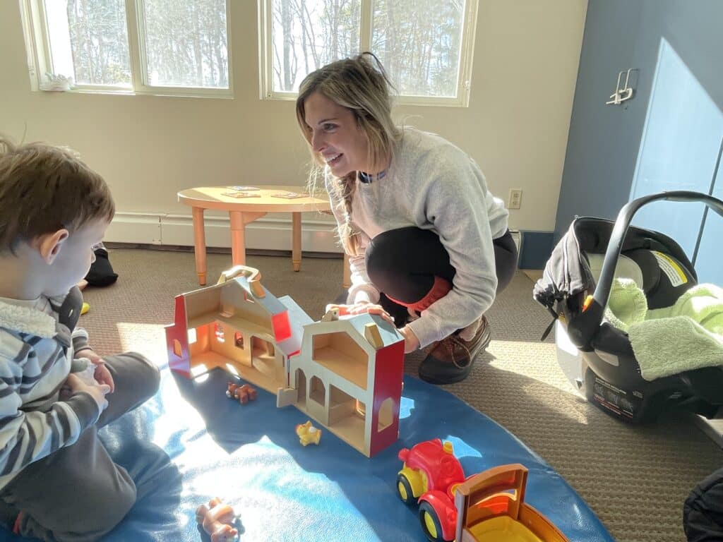 Woman and child playing with a toy barn and animals on a blue mat in a sunlit room.