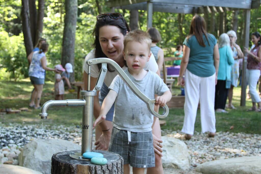 A woman helps a young child use a metal water pump outdoors, with people gathered in the background.
