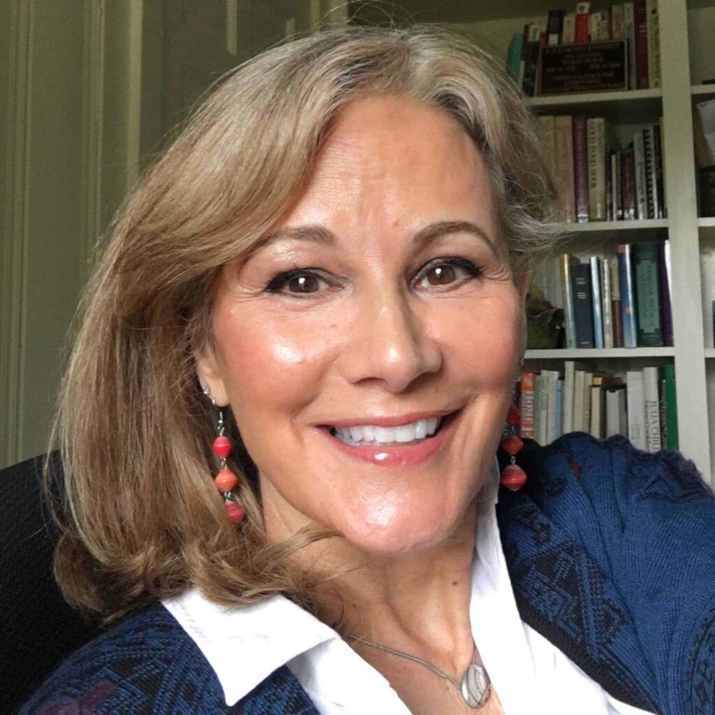Smiling woman with shoulder-length hair, red earrings, and a blue sweater, sitting in front of a bookshelf.