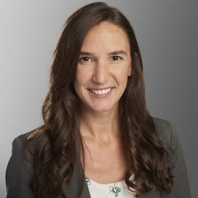 Smiling woman with long brown hair in a gray blazer and floral blouse, posing against a gray background.