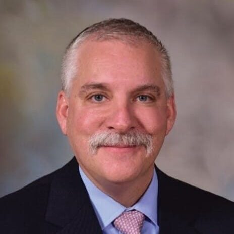 A man with short gray hair, a mustache, and a suit with a light pink tie, smiling at the camera.