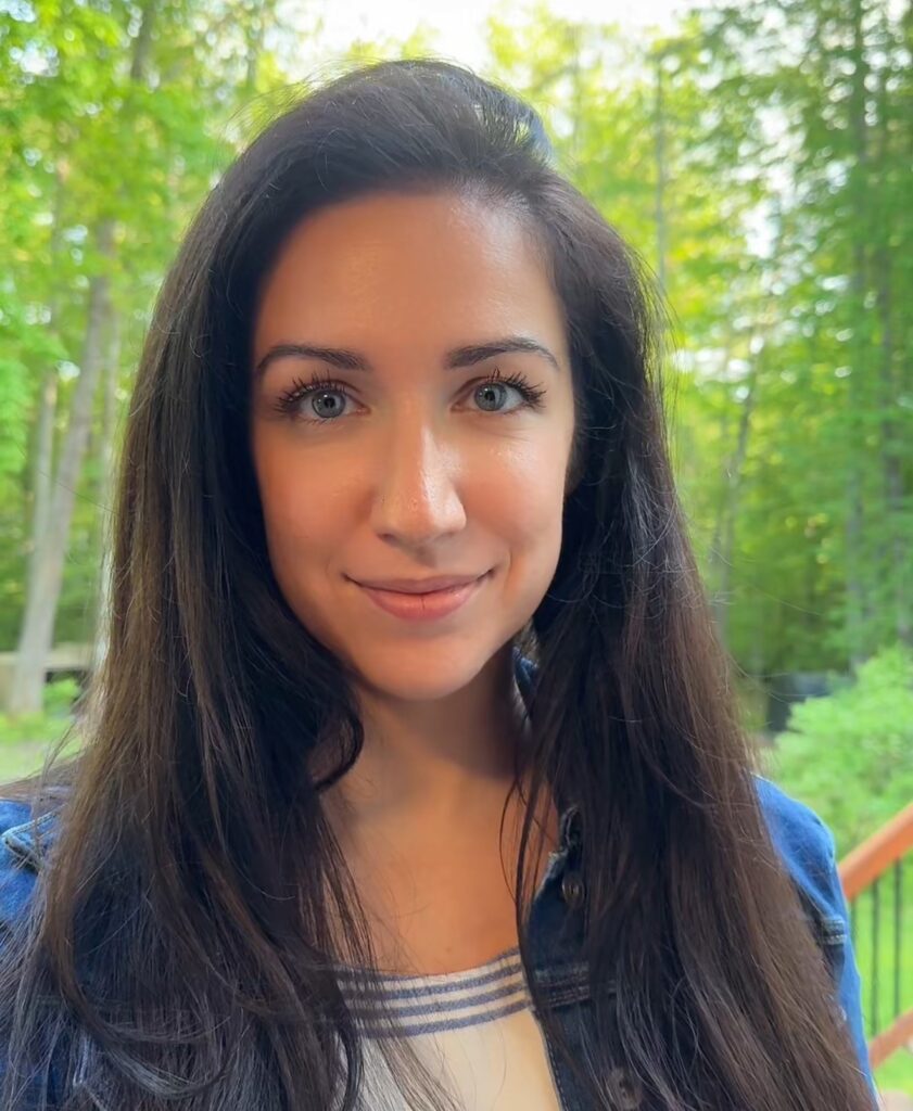 A woman with long dark hair smiles outdoors with green trees in the background.