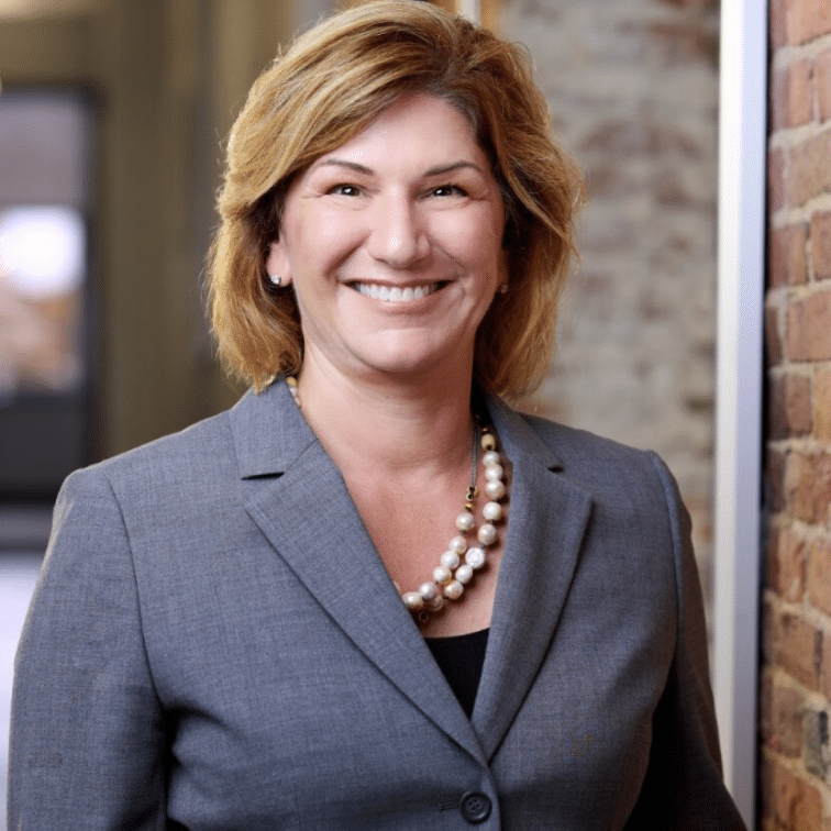 Smiling woman in a gray blazer and pearl necklace stands by a brick wall in an office setting.