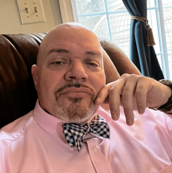 man with a goatee in a pink shirt and checkered bow tie, relaxing on a brown chair by a window.