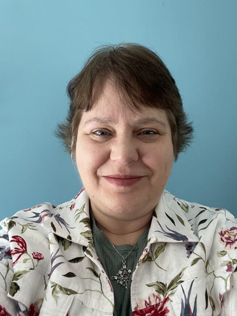 Smiling woman with short brown hair, wearing a floral shirt, in front of a blue wall.