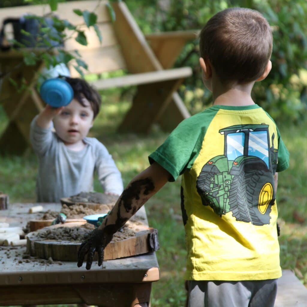 Two young boys play with mud outdoors; one has a muddy arm and the other holds up a blue cup.