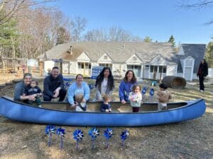 Waypoint staff and families sit in a blue canoe outside Waypoint's Stratham Family Resource Center, celebrating Child Abuse Prevention Month with blue pinwheels — a national symbol of child abuse prevention.