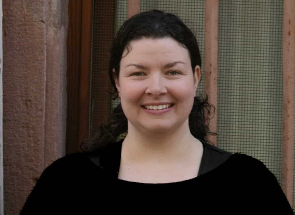 A woman with dark curly hair smiles while standing in front of a window with a metal screen.