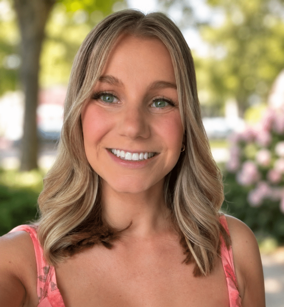 Smiling woman with blonde hair in a pink dress standing outdoors, greenery and flowers blurred in the background.