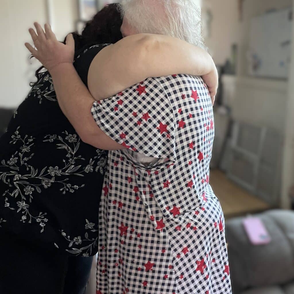 Two people, seen from behind, hugging each other closely in a home setting.