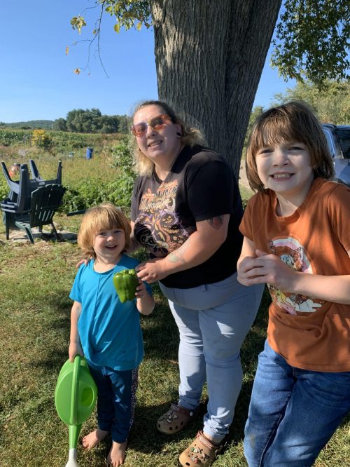 A smiling adult and two children stand outside by a tree, one child holding a green bell pepper.