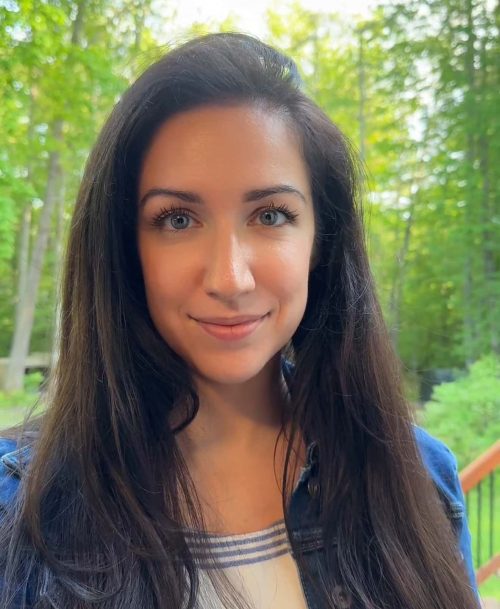 A woman with long dark hair smiles outdoors with green trees in the background.
