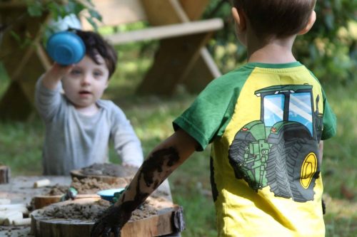 Two young boys play with mud outdoors; one has a muddy arm and the other holds up a blue cup.