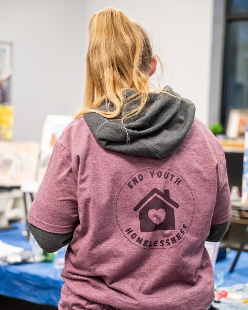 Person with long hair in a ponytail wears a shirt reading End Youth Homelessness with a house and heart logo.