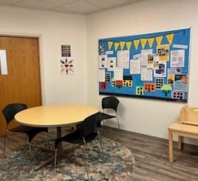 Small meeting room with a round table, four chairs, and a bulletin board covered with papers and decorations.