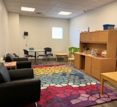 Colorful rug, black chairs, tables, and wooden shelves in a bright, organized room with white walls and ceiling lights.