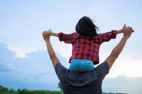 Adult carrying child on shoulders outdoors, both holding hands, child wearing red plaid shirt, sky in background.